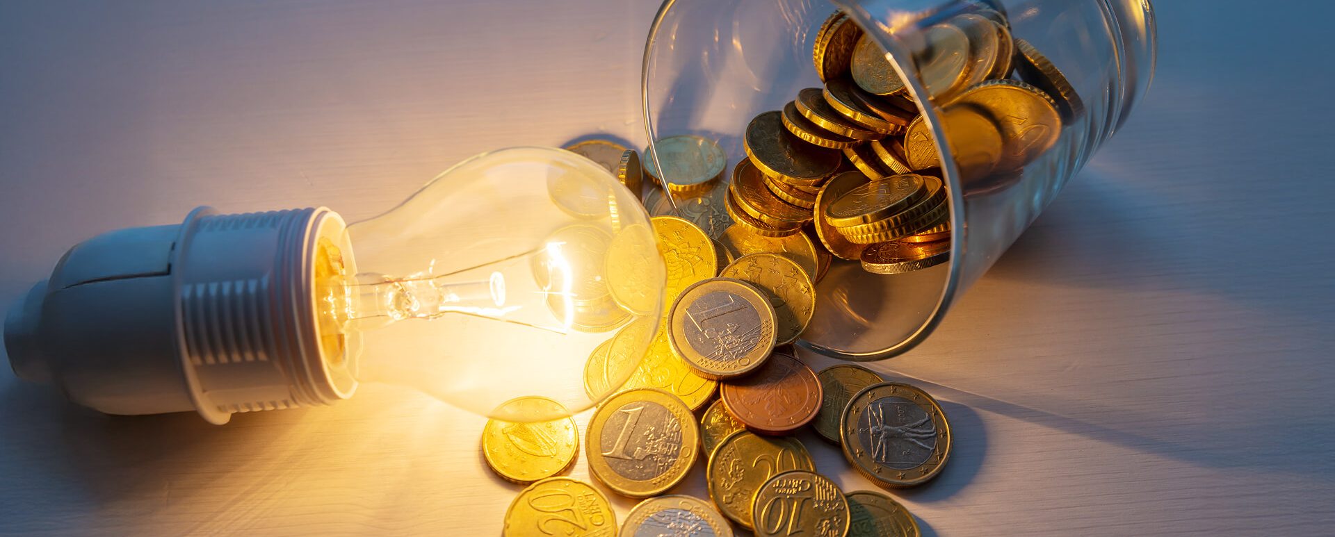 Ein Glas in dem sich Münzen befinden daneben eine leuchtende Glühbirne