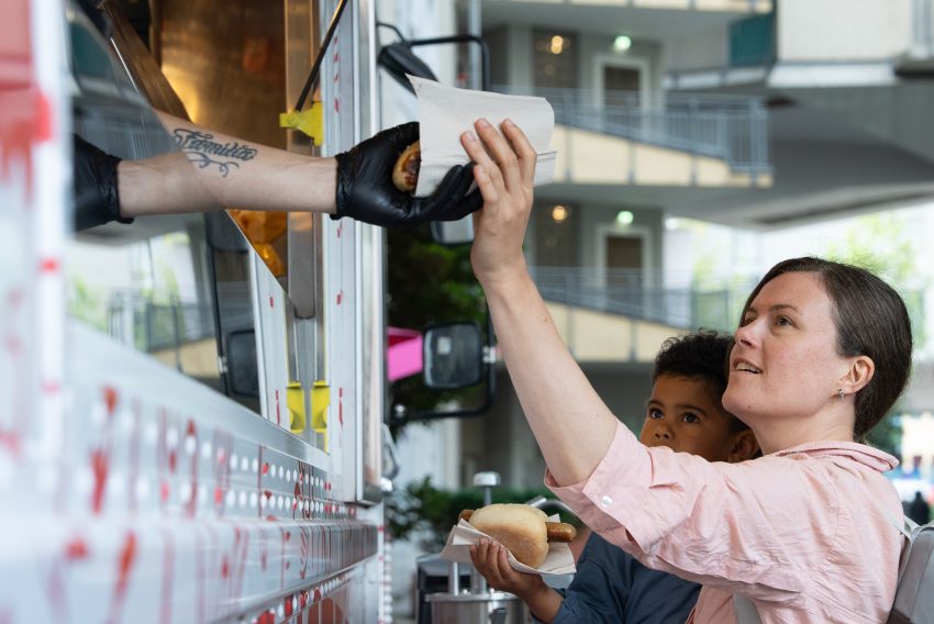 Hand mit schwarzem Handschuh reicht Bratwurst in Papier an Person in rosa Hemd vor Imbisswagen