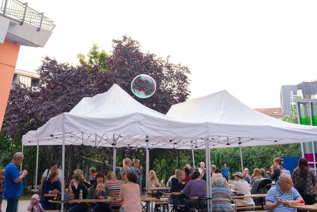 Mehrere Personen sitzen unter zwei weißen Pavillons auf Holzbänken und -tischen im Freien, im Hintergrund Bäume und Gebäude.