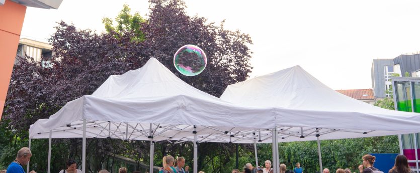 Mehrere Personen sitzen unter zwei weißen Pavillons auf Holzbänken und -tischen im Freien, im Hintergrund Bäume und Gebäude.