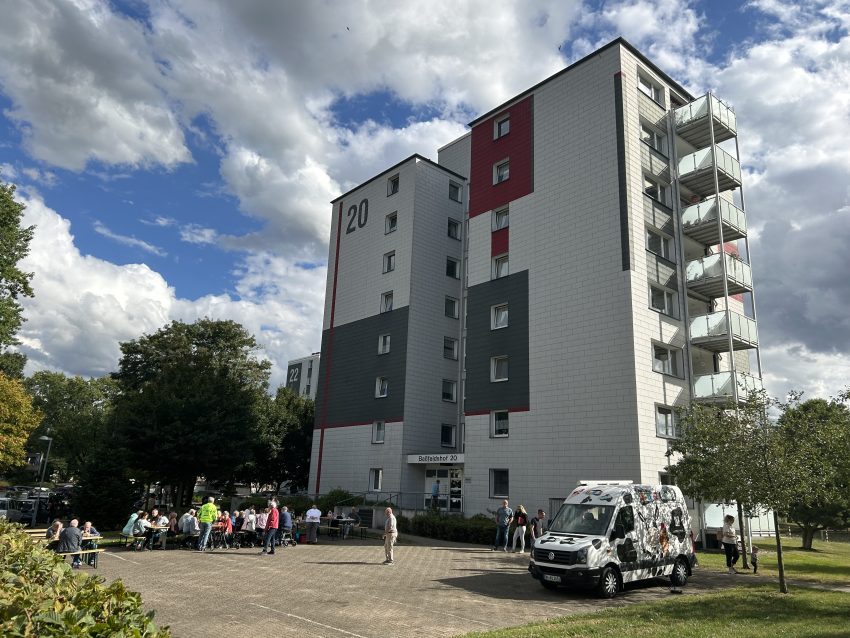Mehrstöckiges Wohnhaus, davor Menschen auf Bänken und ein weißer Kleintransporter auf dem Hof.