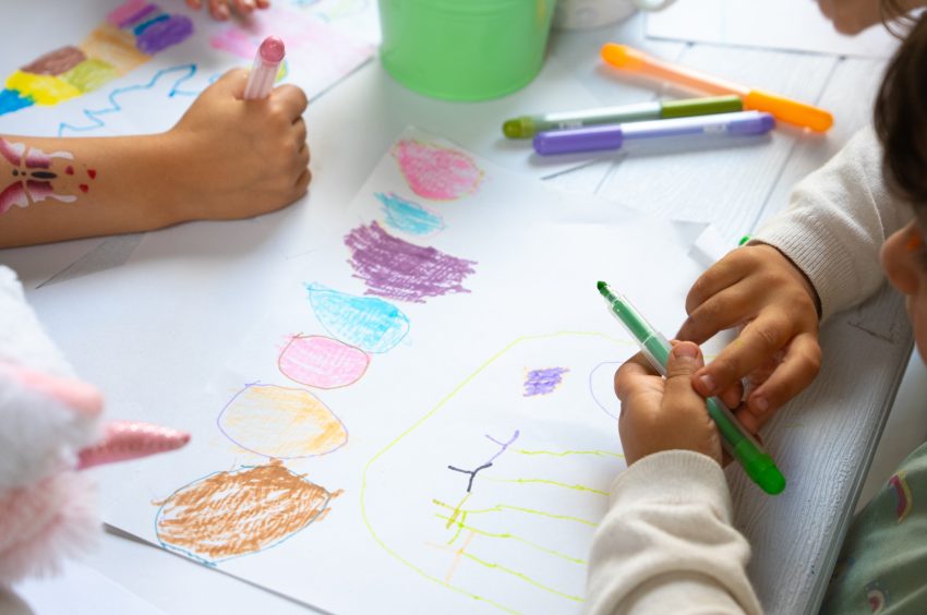 Kinderhände zeichnen mit bunten Filzstiften auf weißem Papier bunte Formen und Linien.