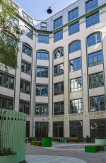 Innenhof eines mehrstöckigen Gebäudes mit weißen Fassaden, vielen Fenstern und grünen Sitzgelegenheiten auf dem gepflasterten Boden.