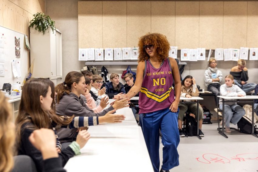 Person mit lockigem Haar und Sporttrikot geht an einer Tischreihe mit mehreren sitzenden Personen vorbei und gibt ihnen High-Fives