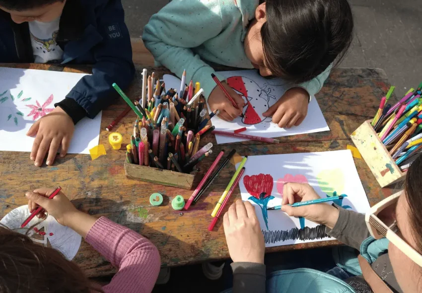 Mehrere Kinder sitzen an einem Holztisch und malen mit Buntstiften auf Papier, auf dem Blumen und andere bunte Motive zu sehen sind.