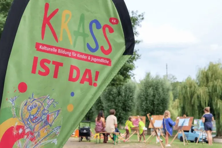 Grüne Fahne mit der Aufschrift 'KRASS IST DA!' vor einer Gruppe von Kindern, die im Freien an Staffeleien malen