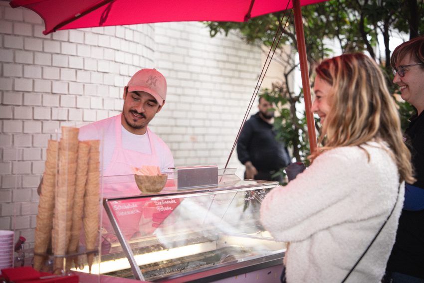 Verkäufer mit rosa Schürze hinter Eisvitrine mit Eiswaffeln, Kundin mit weißem Pullover davor, roter Sonnenschirm über dem Stand.