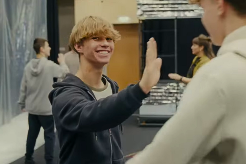 Junge hebt die Hand zum Abklatschen vor einer Person in weißem Oberteil in einem Raum mit weiteren Personen im Hintergrund