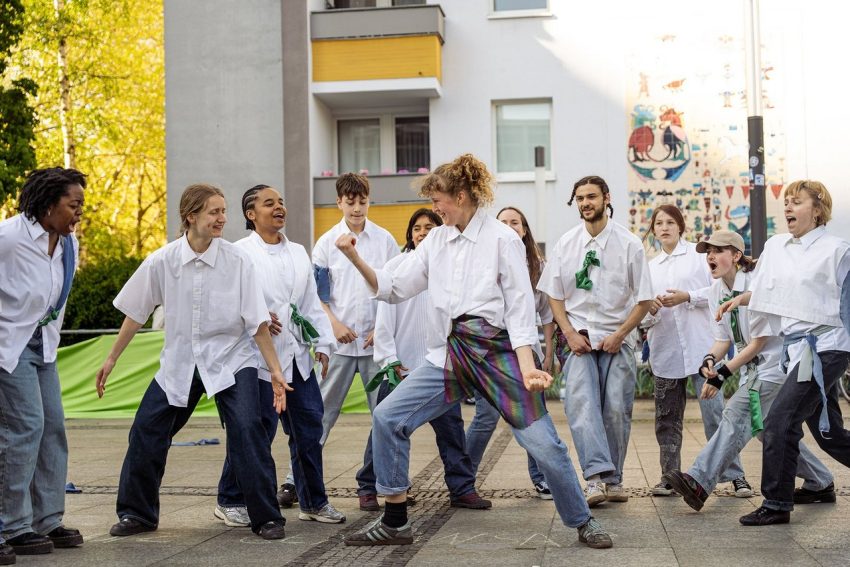 Gruppe von Jugendlichen in weißen Hemden und Jeans steht auf einem Hof vor einem Wohnhaus und tanzen