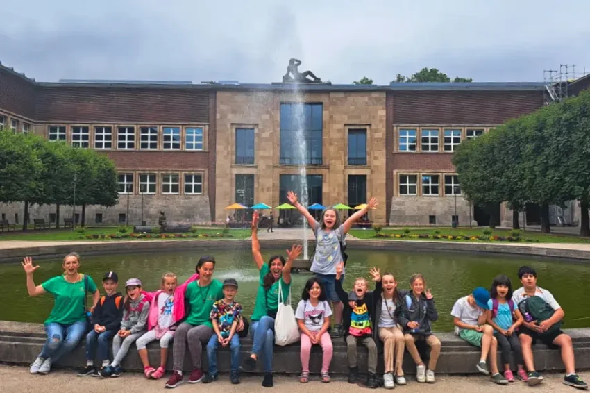 Gruppe von Kindern und Jugendlichen sitzt und steht vor einem großen Gebäude mit Brunnen und Bäumen